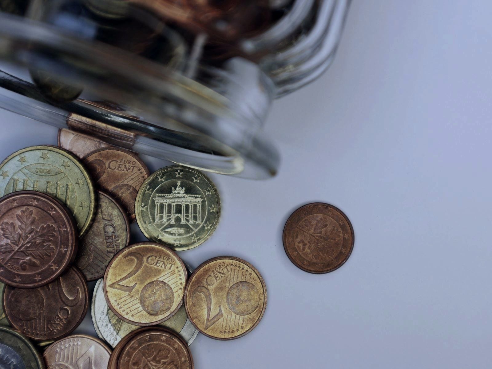 Euro coins of several values spilling from a glass jar onto a white surface.