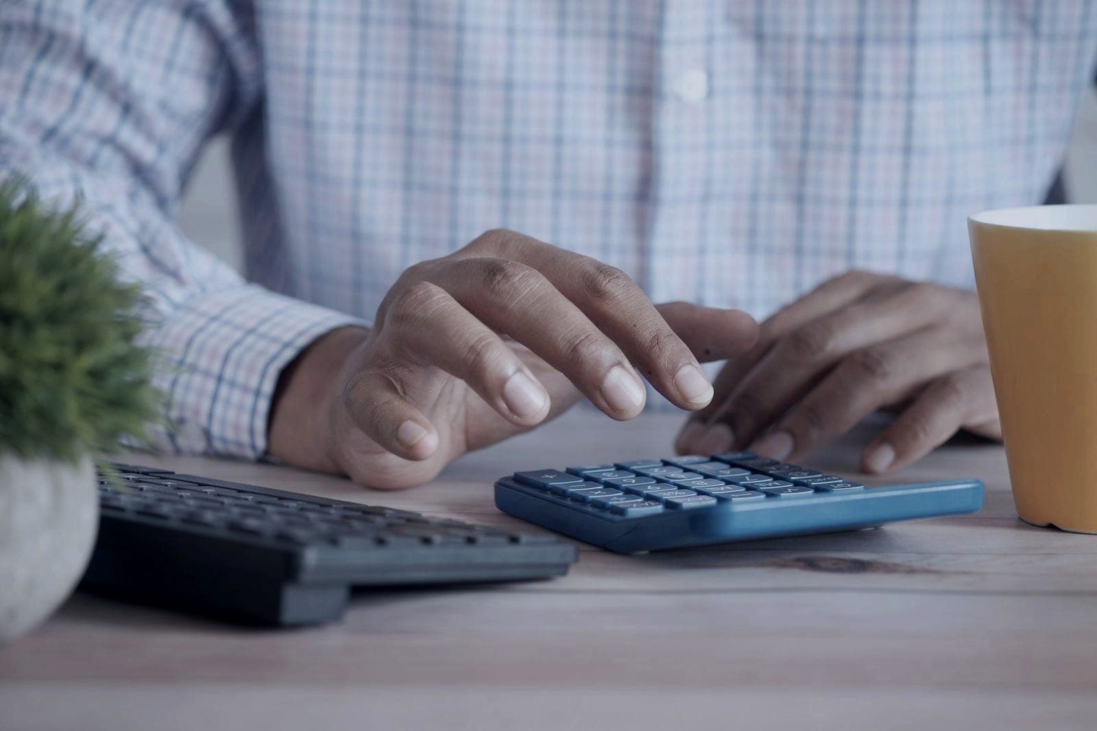Mãos a usar uma calculadora azul numa secretária, ao lado de um teclado e de uma caneca.