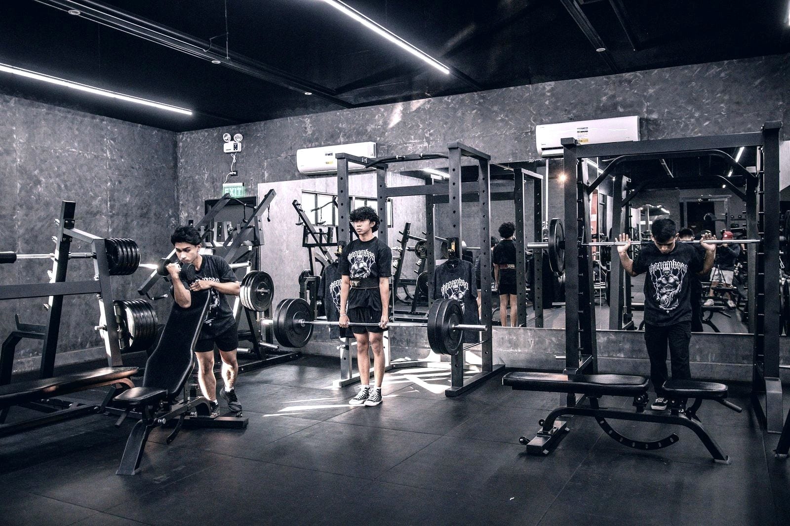 People training with barbells in squat racks in a dark-walled training room.