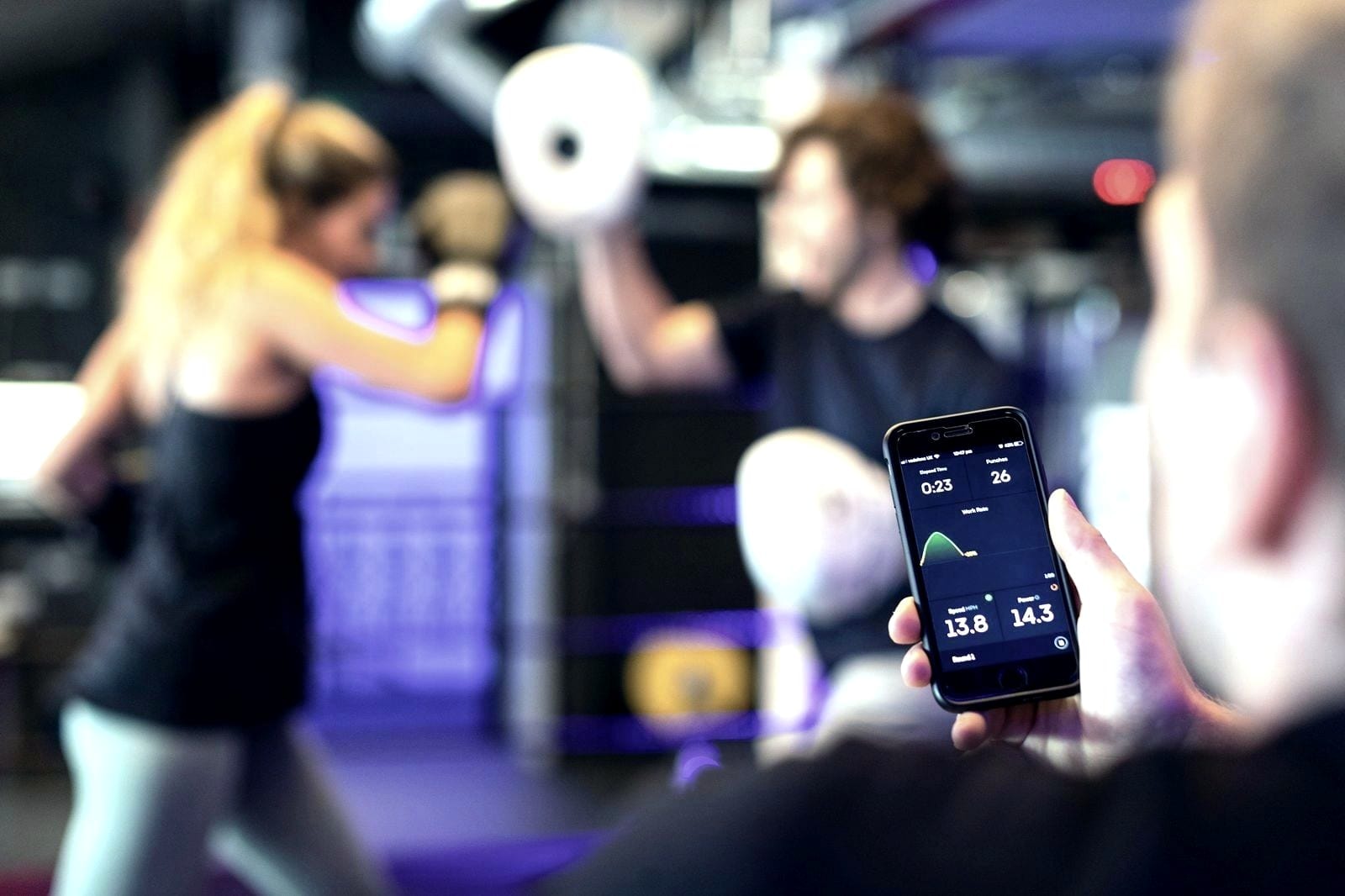 Hand holding a phone with workout data, with a class in progress blurred behind it.