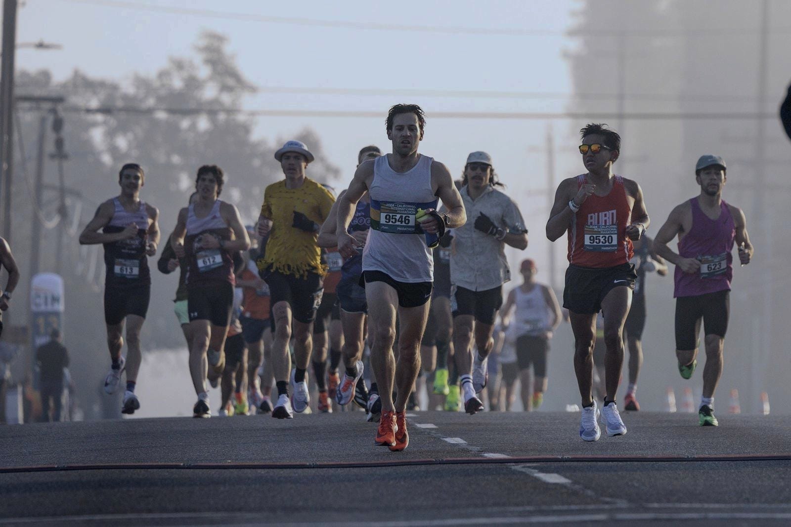 A group of runners wearing race bibs on a road, on a misty morning.
