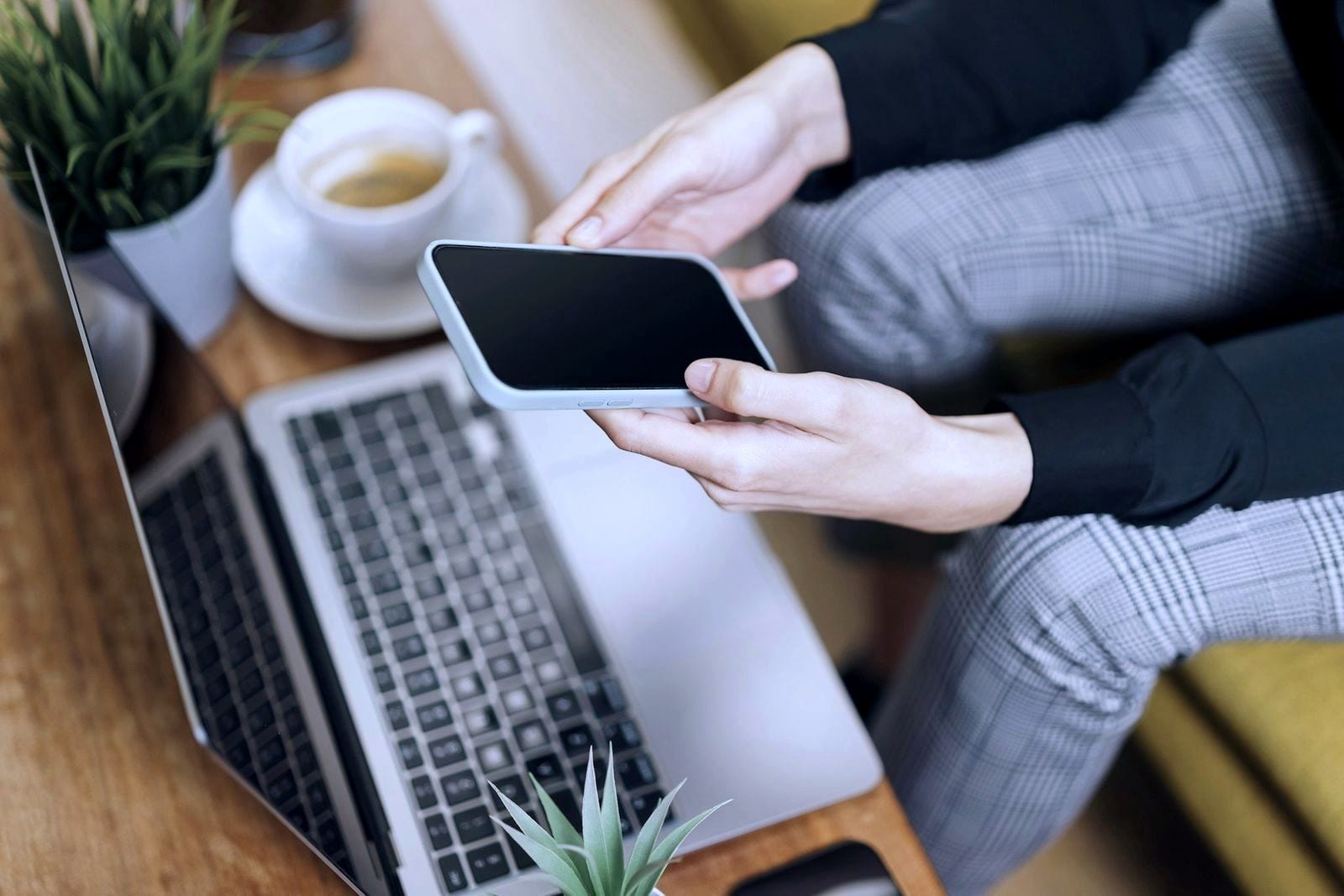 Hands holding a phone above an open laptop on a wooden table.